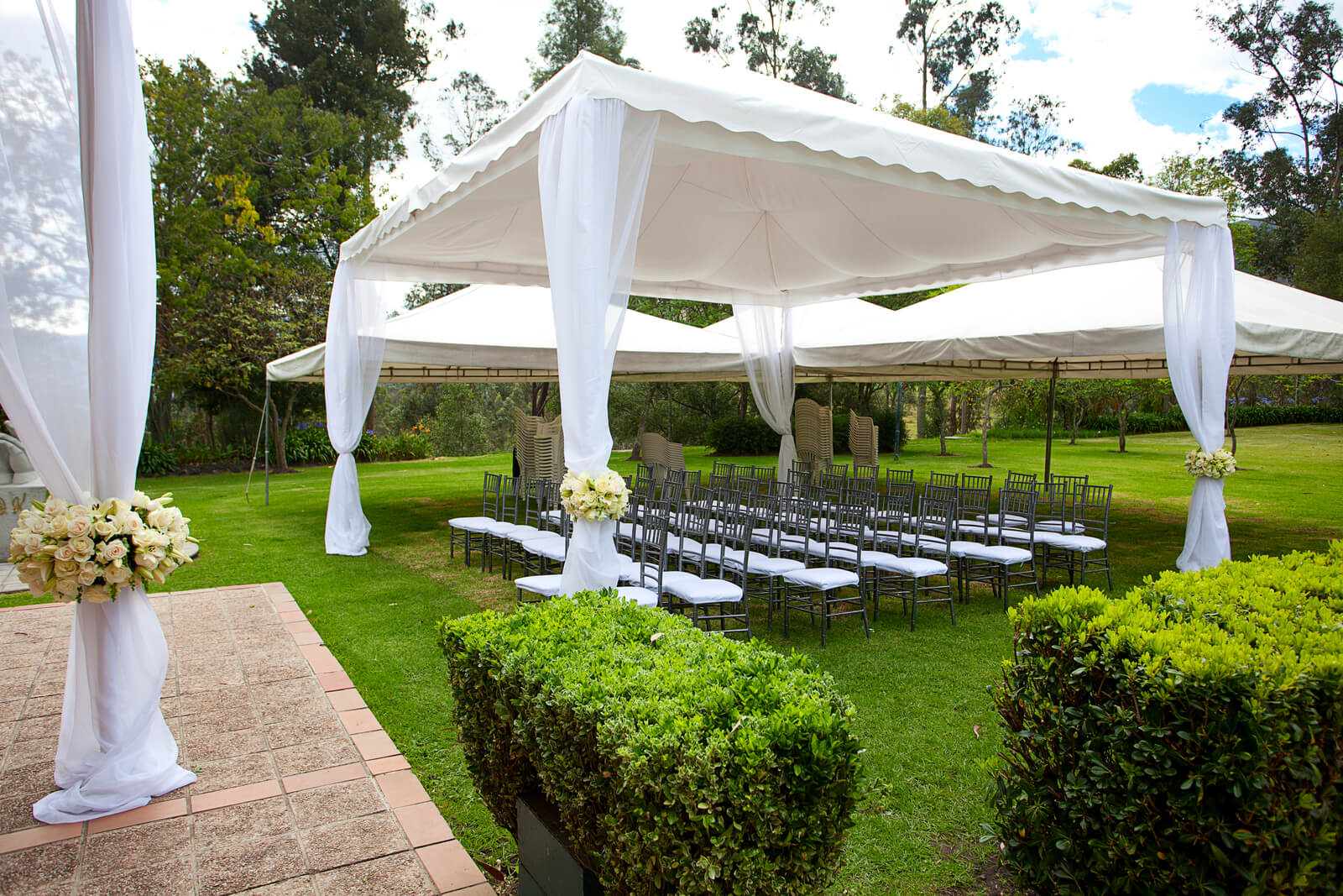 Lawn with surrounding trees and bushes, prepared for a wedding with white drapes, white and grey lawn chairs and small festival tents.