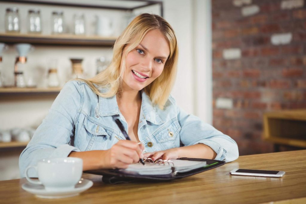 Smiling woman writing things down in a binder, sitting in a coffeeplace with her phone and a cup of coffee on the table.