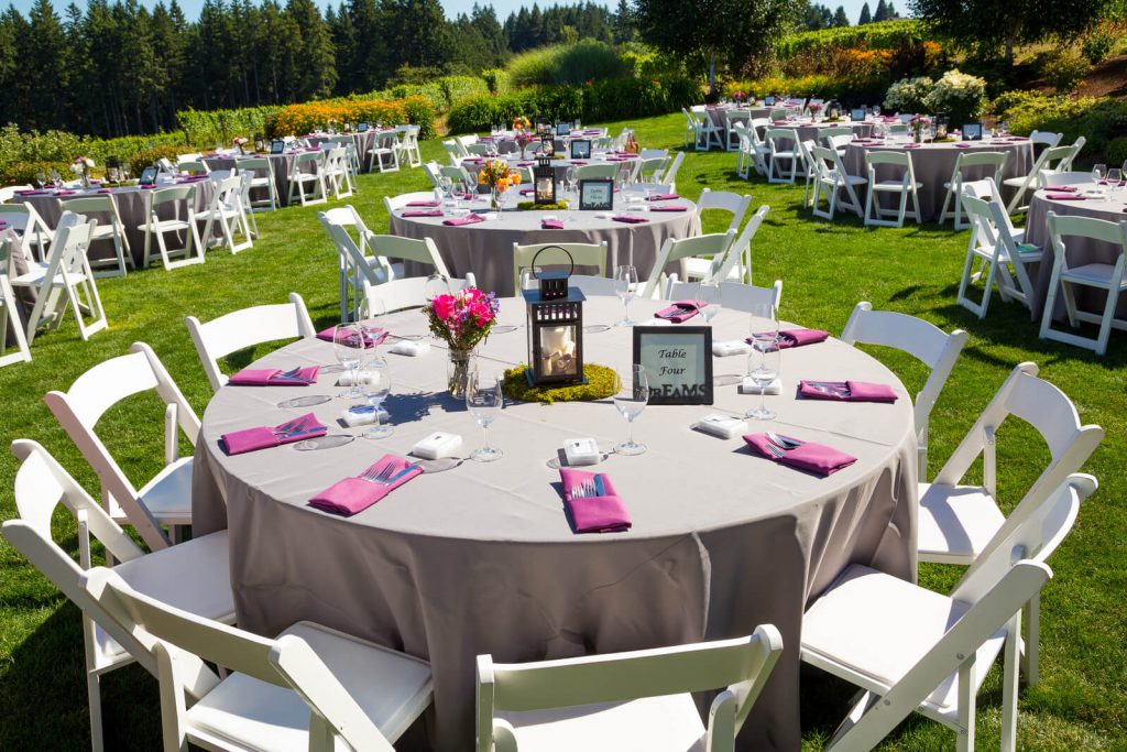 Rented tables and chairs at a wedding. All setup with cutlery, purple and grey finishes on a green lawn surrounded by nature